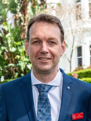 A man in a tie, white shirt and blue jacket smiles. He is outdoors with a building and tree in the background. 